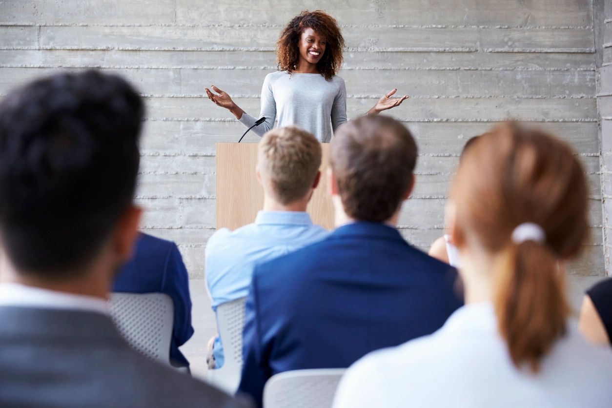 A Woman In Light Blue Talking Over A Podium